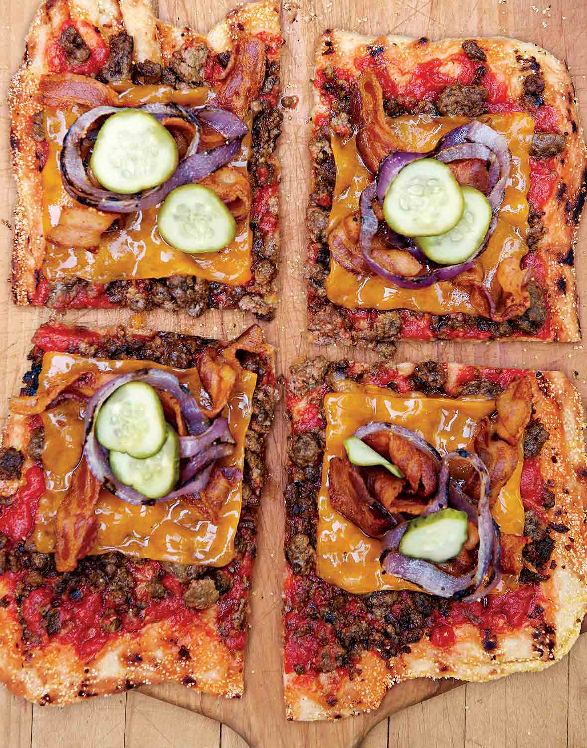 A bacon cheeseburger pizza topped with beef, bacon, pickles, cheese, and red onion, cut into four squares on a wooden pizza peel.