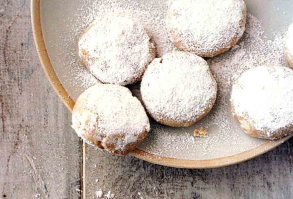 A bowl of cardamom and cashew cookies with one broken cookie beside the bowl.