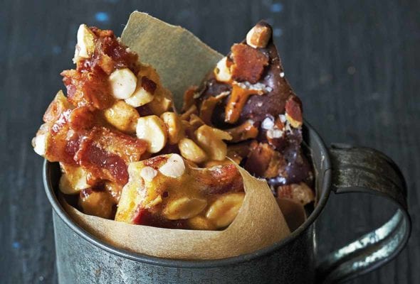 Tin cup lined with brown paper and filled with peanut brittle studded with bacon on blue wood