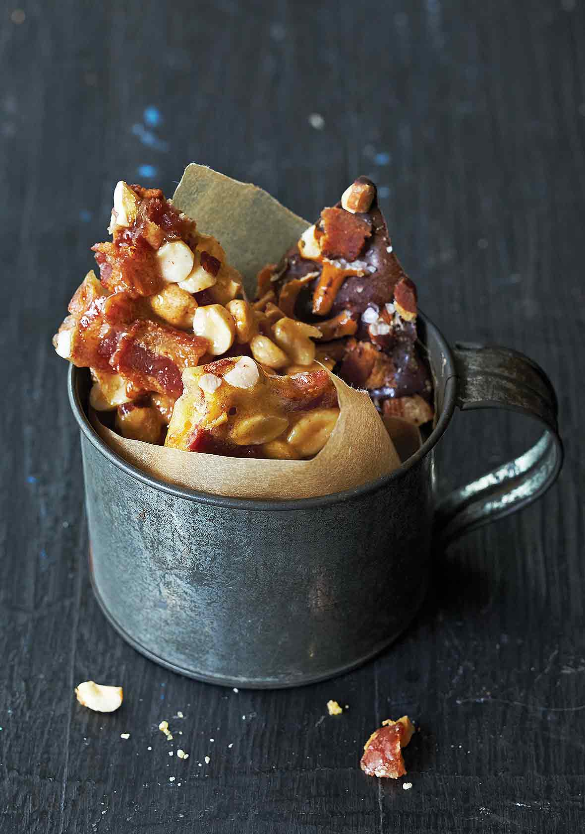 Tin cup lined with brown paper and filled with peanut brittle studded with bacon on blue wood