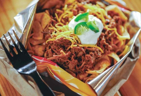 Ripped open bag of Fritos, on top is beef chili, shredded cheese, sour cream, and sliced peppers