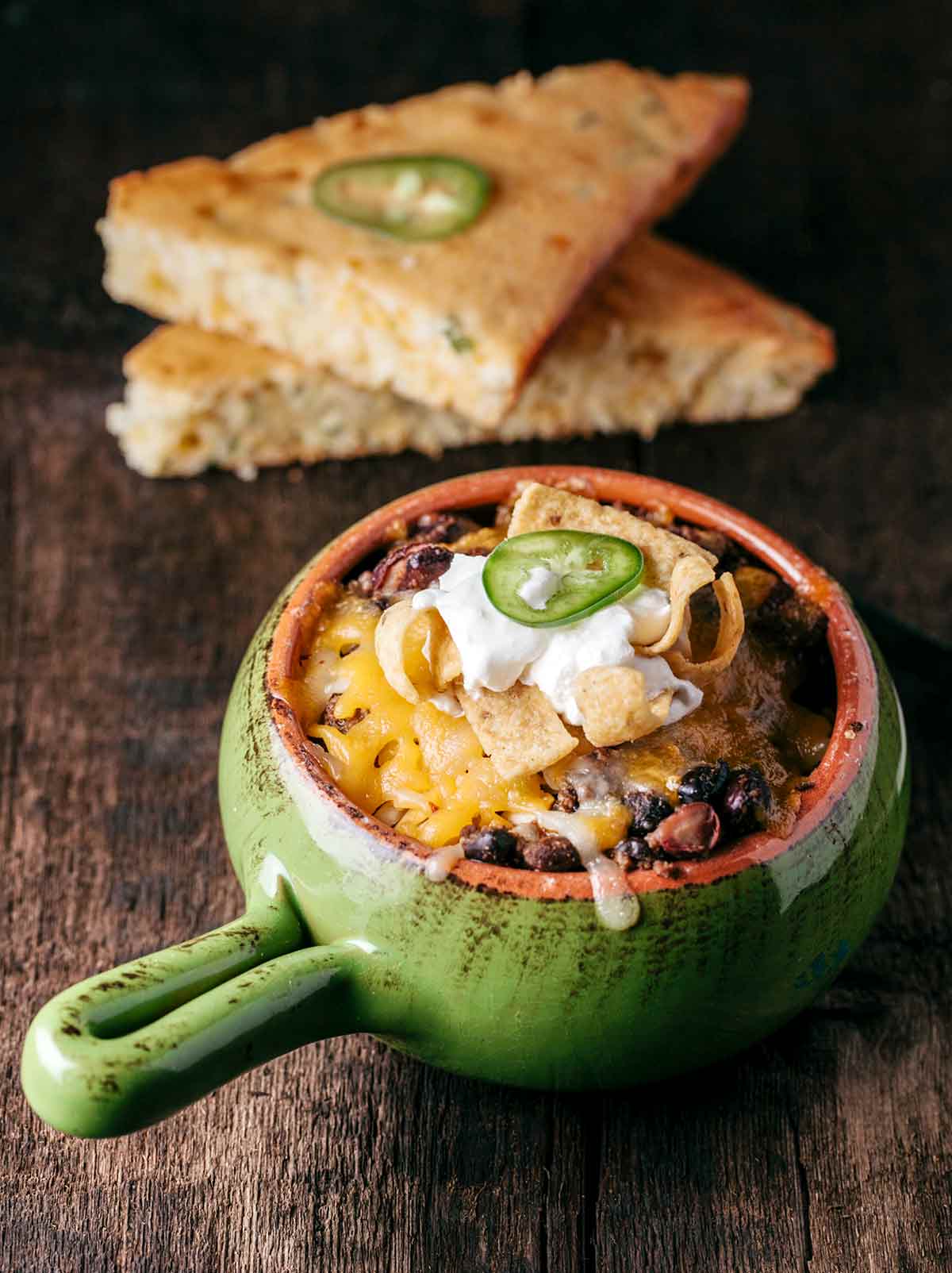 Frito pie in a green enamel bowl with a handle with two pieces of jalapeno bread in the background.