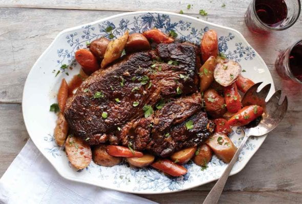 A pot roast with potatoes and vegetables on a decorative platter with a serving fork on the platter and a napkin on the side.