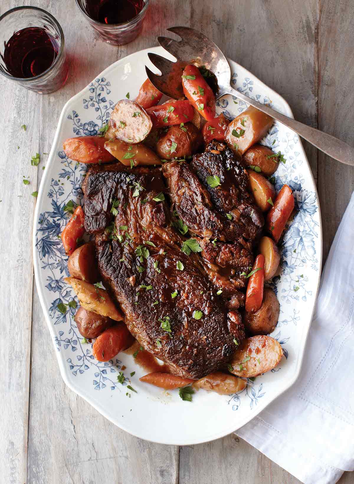 A pot roast with potatoes and vegetables on a decorative platter with a serving fork on the platter and a napkin on the side.