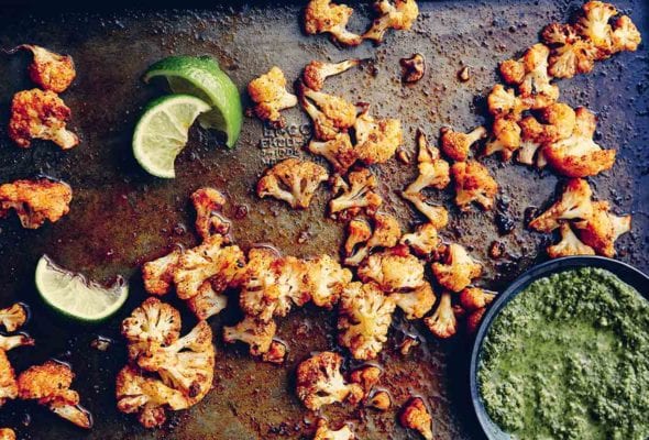 Florets of spicy roasted cauliflower on a baking sheet with squeezed lime wedges and a bowl of green sauce beside them.