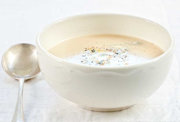 A white bowl of cauliflower soup on a white linen cloth with a spoon resting beside it.
