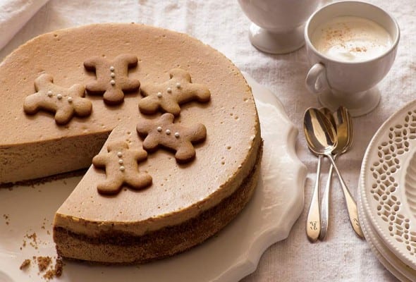 A gingerbread cheesecake with one slice missing on a decorative plate with spoons and espresso cups on the side