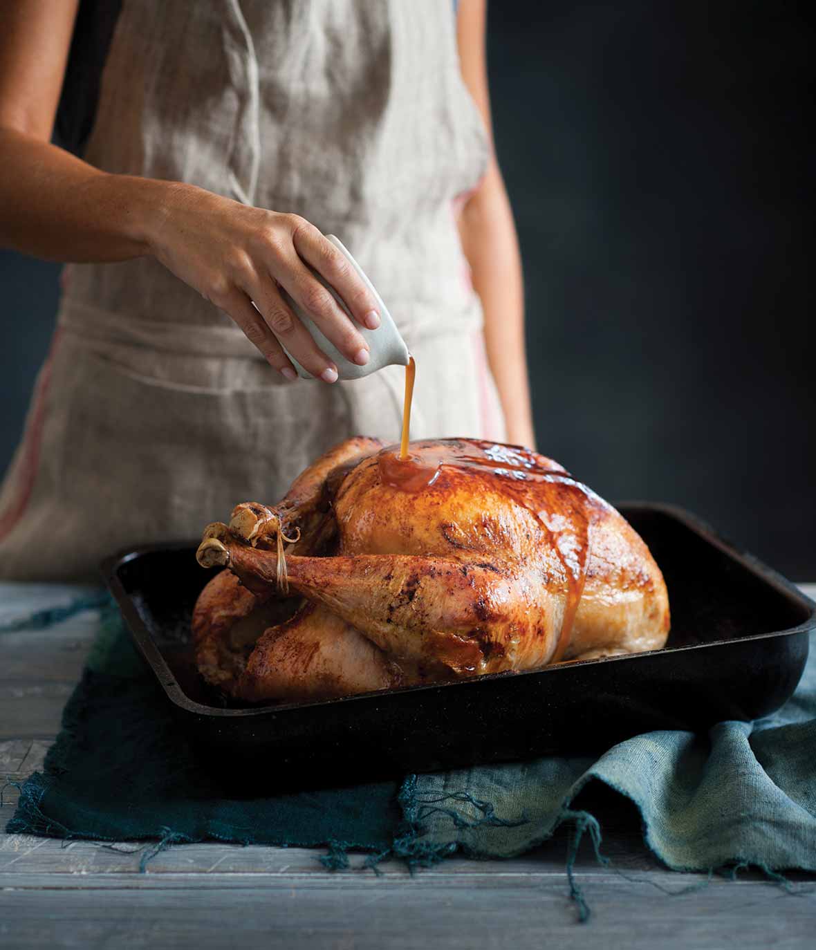 A woman pouring maple syrup over a maple glazed turkey in a black roasting pan.