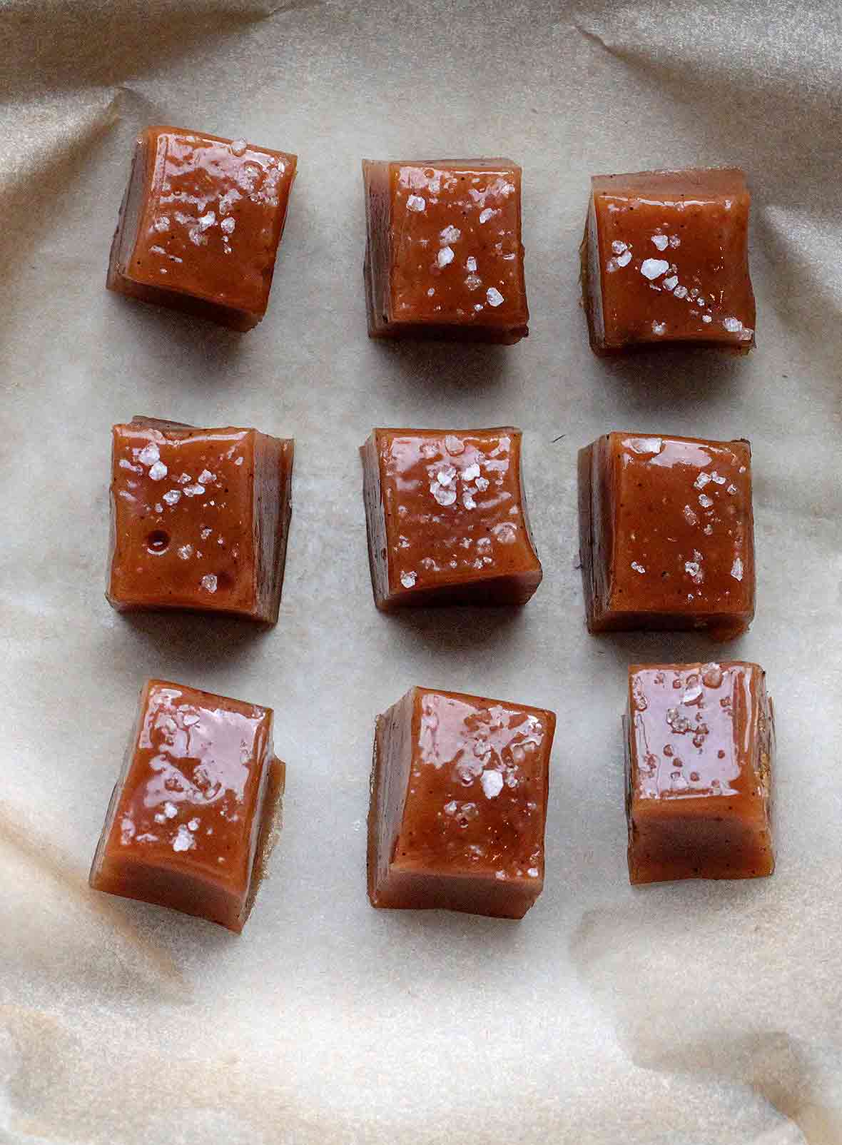 Six square caramels topped with sea salt sitting on a sheet of parchment.