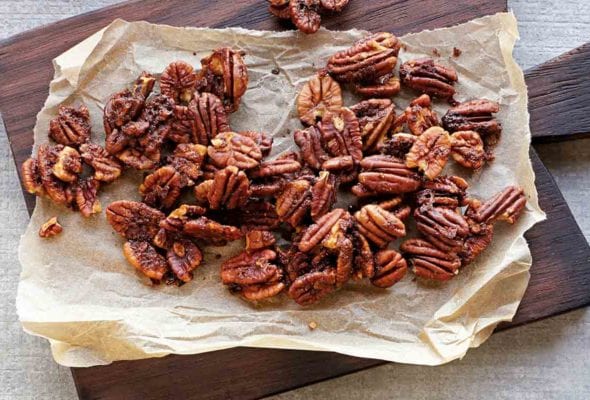 Baked spiced pecans on a sheet of parchment that is resting on a wooden serving board.