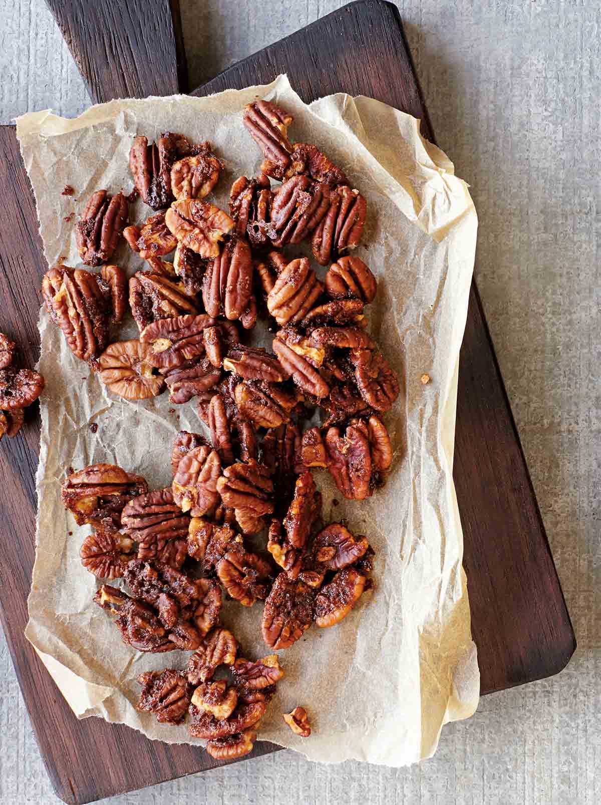 Baked spiced pecans on a sheet of parchment that is resting on a wooden serving board.