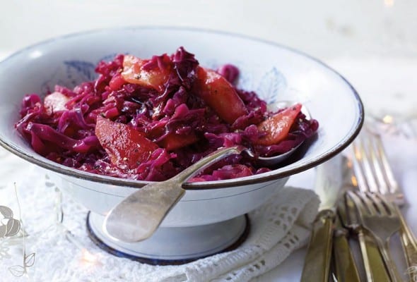 A white bowl filled with spiced red cabbage set on white linens with a spoon resting inside.