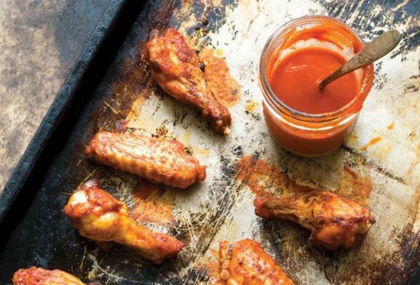 A baking sheet with 6 spicy baked chicken wings glazed with hot sauce, a jar of sauce nearby