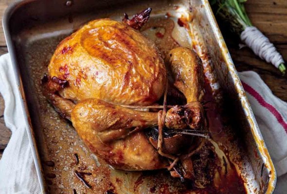 A perfect roast chicken in a metal roasting pan that is resting on a kitchen towel, with a bunch of herbs beside it.