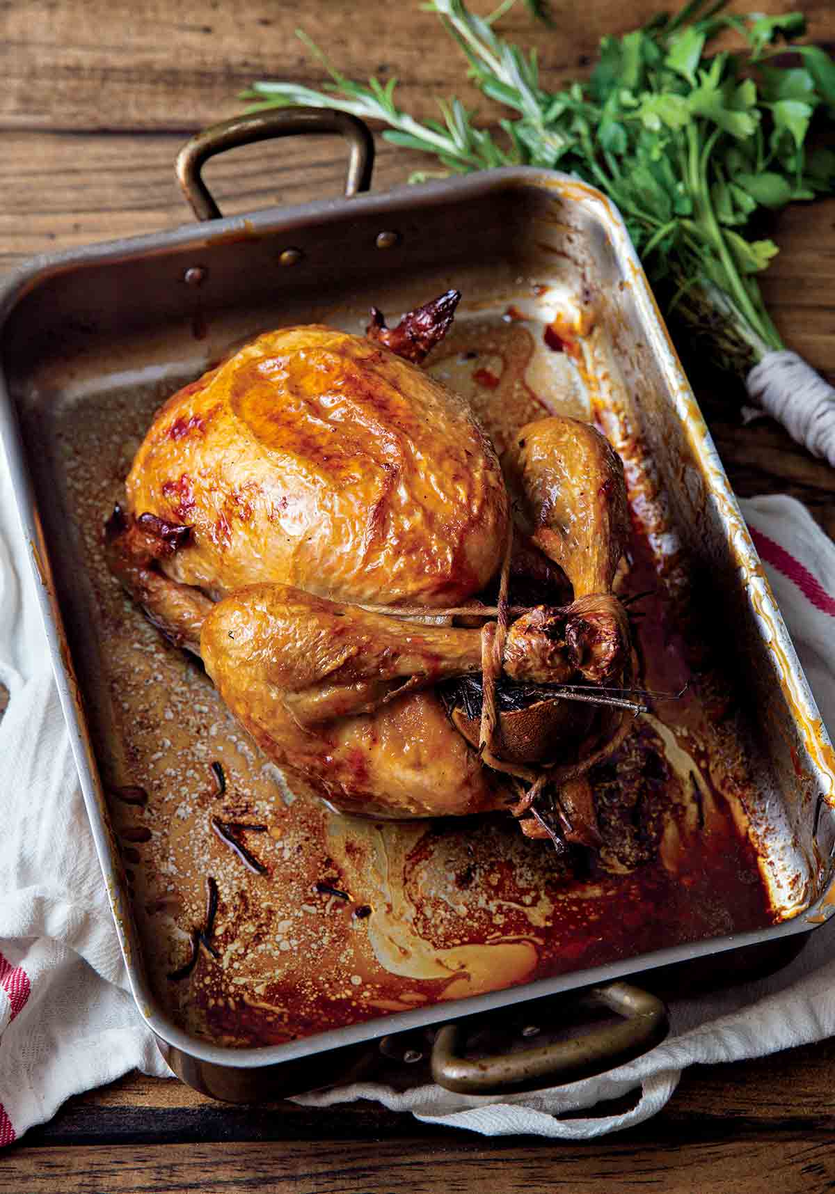 A perfect roast chicken in a metal roasting pan that is resting on a kitchen towel, with a bunch of herbs beside it.