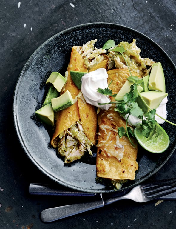 Two chicken enchiladas with green sauce in a black shallow bowl with cubed avocado, sour cream, cilantro, and a lime half.