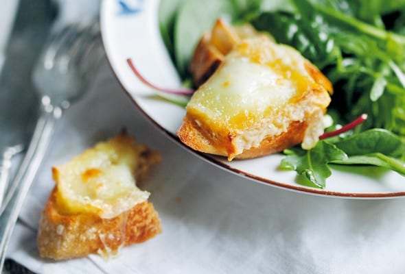A bowl filled with baby greens on a white linen napkin. Part of a goat cheese crostini is on the edge of the bowl and the other part on the napkin