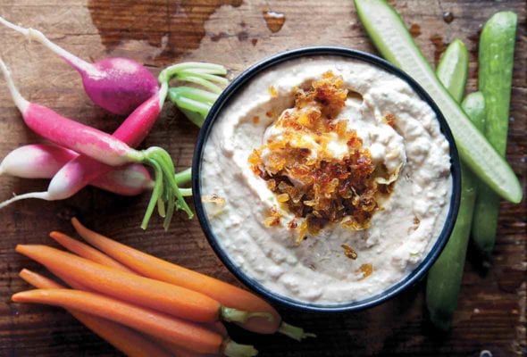 Bowl of onion dip topped with caramelized onions, nearby carrots, radishes, cucumbers on a cutting board