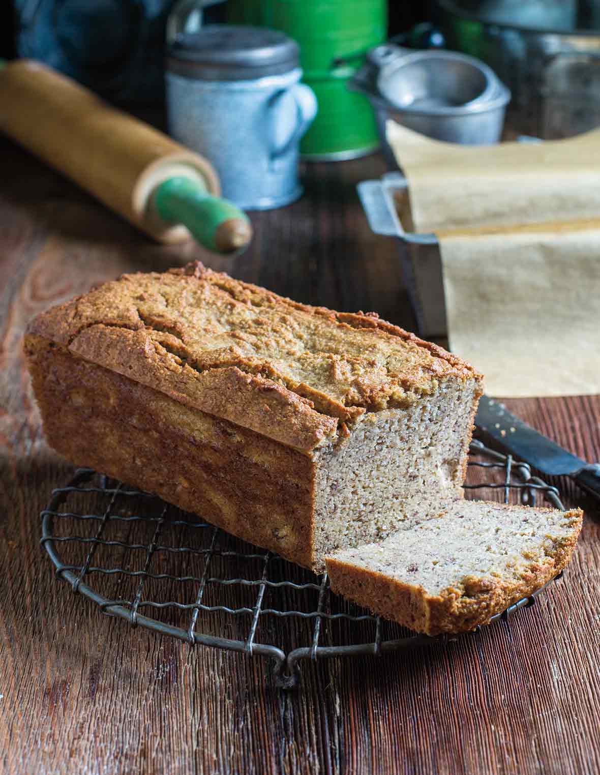 A loaf of paleo banana bread with one slice cut, resting on a wire rack.