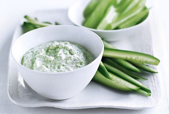 A white platter with sliced celery and cucumber and a white bowl of feta dip on top.