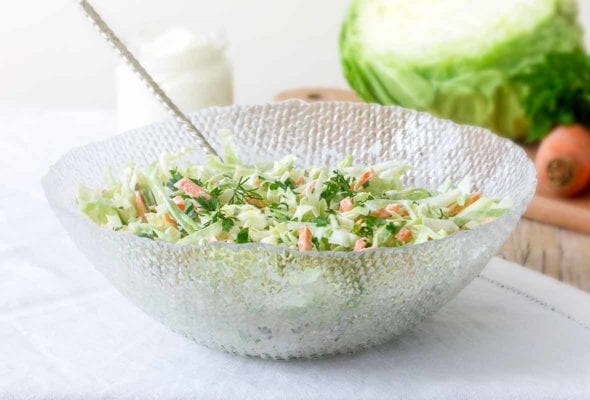 Glass bowl with lime coleslaw--green cabbage, carrots, lime zest, mayo, and parsley--on a tablecloth, cabbage and carrots behind