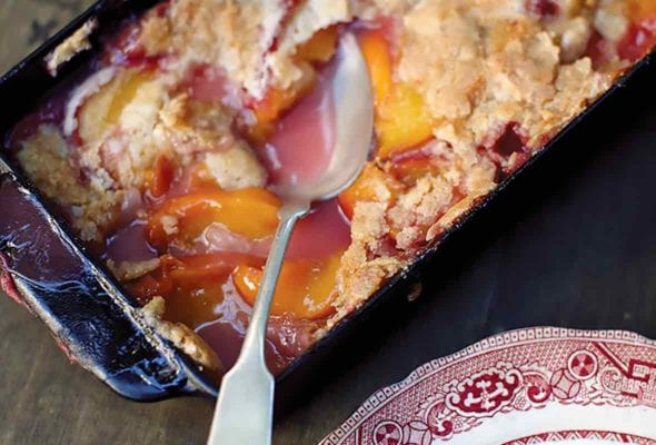 A spoon resting in a glass baking dish of peach cobbler, plus a helping of peach cobbler on a decorative plate.