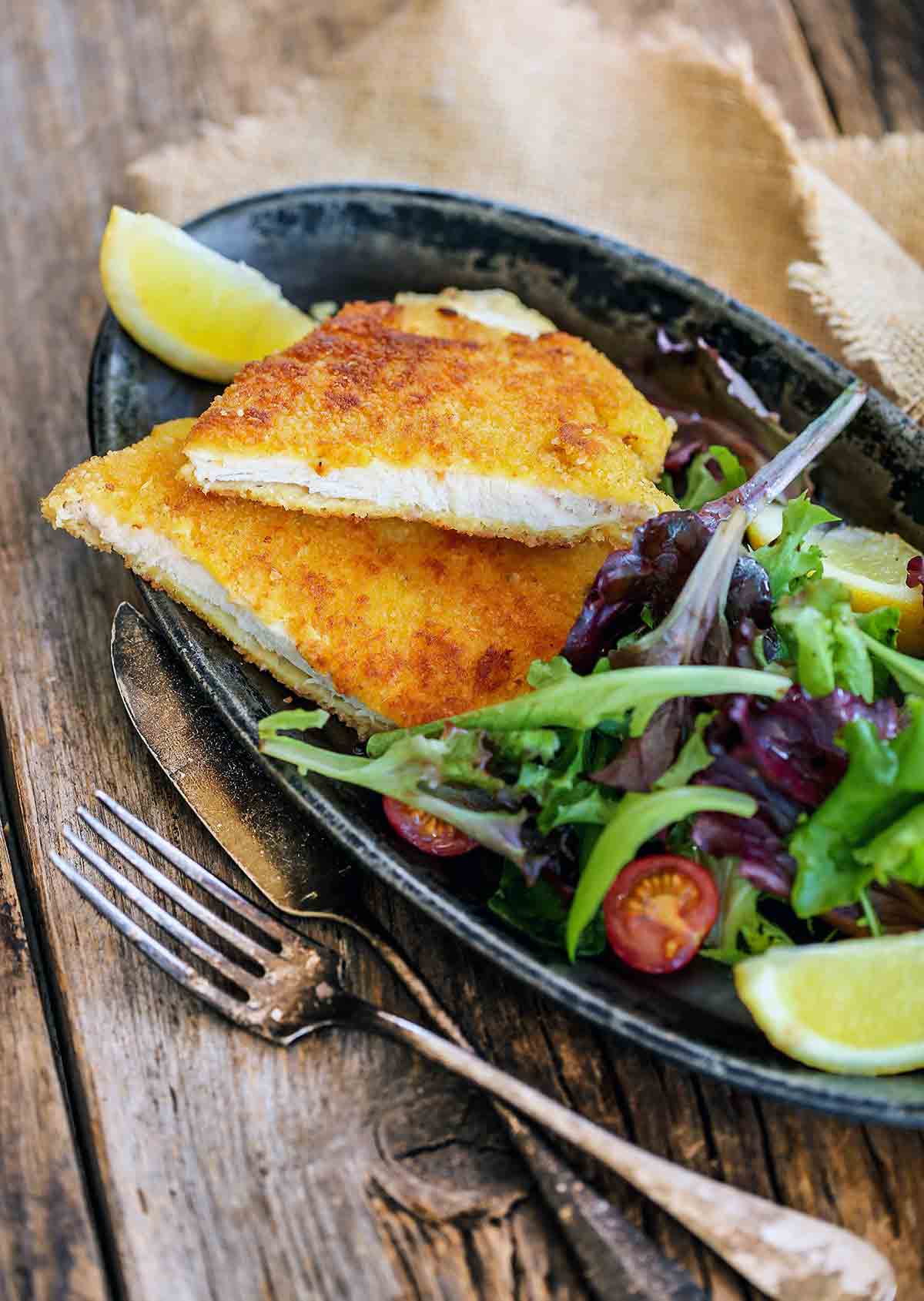 A halved pork Milanese cutlet on a grey oval plate with a lemon wedge and mixed spring greens.