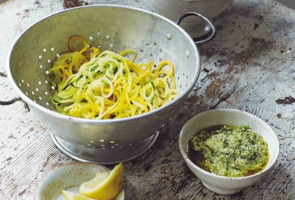 A wooden table with a colander holding veggie spaghetti, a small white bowl of pesto, and a small dish of lemon wedges.