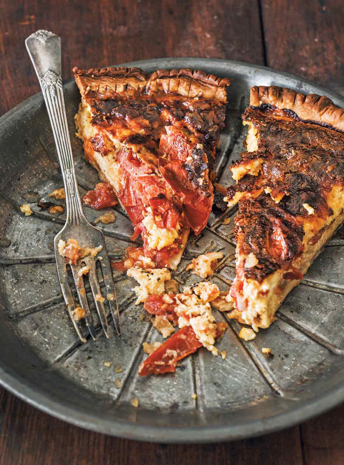 A metal pie plate with two cut slices of roasted tomato quiche and a fork resting beside them.