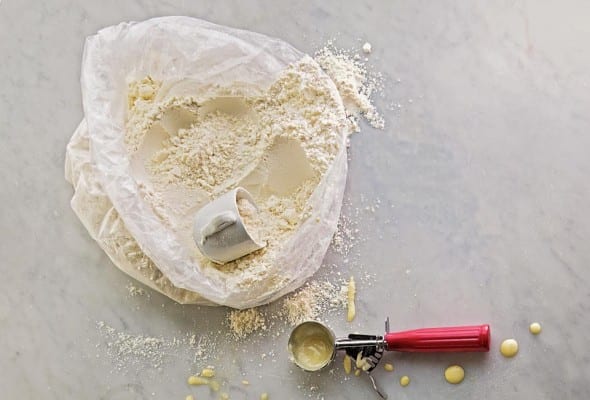 An open plastic bag of homemade pancake mix with a mug for scooping and an ice cream scoop on the side for the batter.