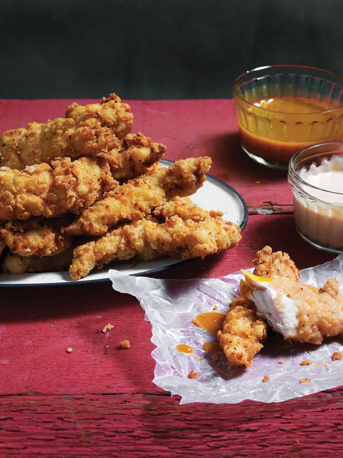 A platter of homemade chicken strips, with two lying beside the platter on a piece of wax paper, and two dipping sauces on the side.