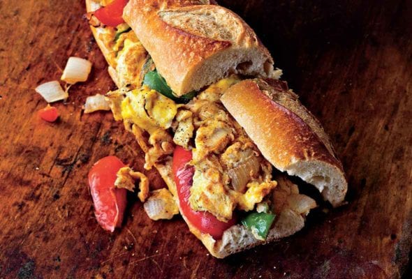 A pepper and egg sandwich on a wooden table with pieces of pepper and onion falling out.