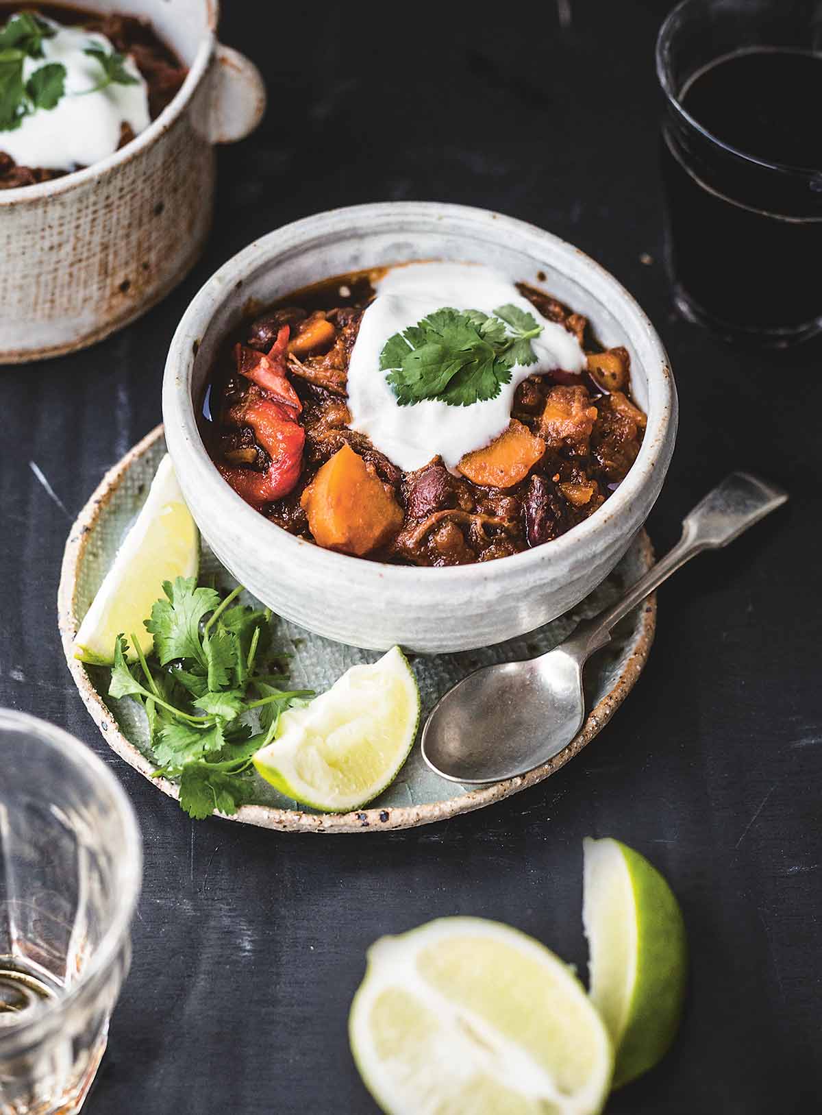 A bowl of shredded beef chili with sweet potatoes on a plate containing a spoon, two lime wedges, and cilantro leaves.