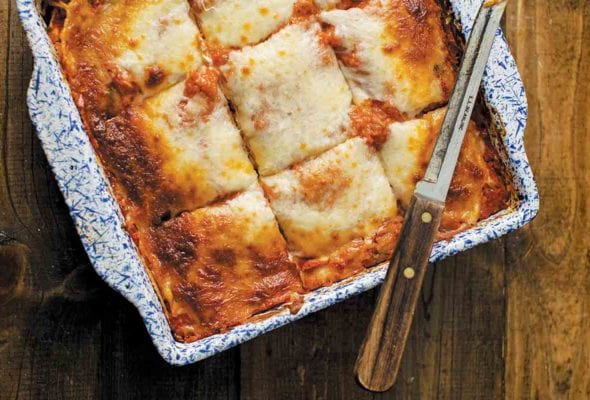 A blue and white square baking dish filled with vegetable lasagna that has been cut into 9 squares with a knife resting on top.
