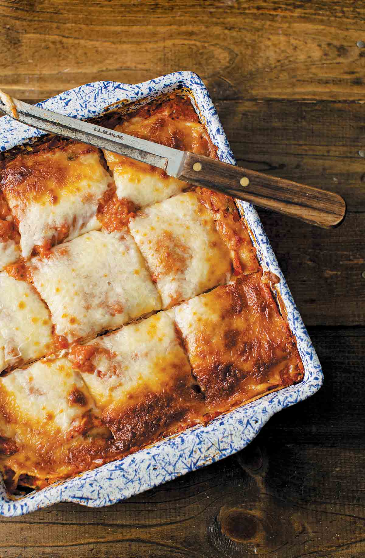A blue and white square baking dish filled with vegetable lasagna that has been cut into 9 squares with a knife resting on top.