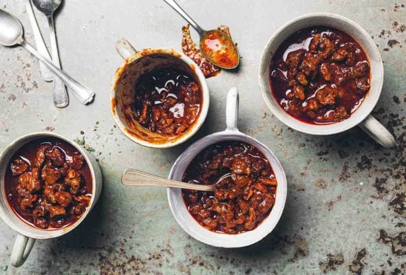 Four mugs filled with chili con carne with one spoon resting inside a mug and four others laying beside the mugs.