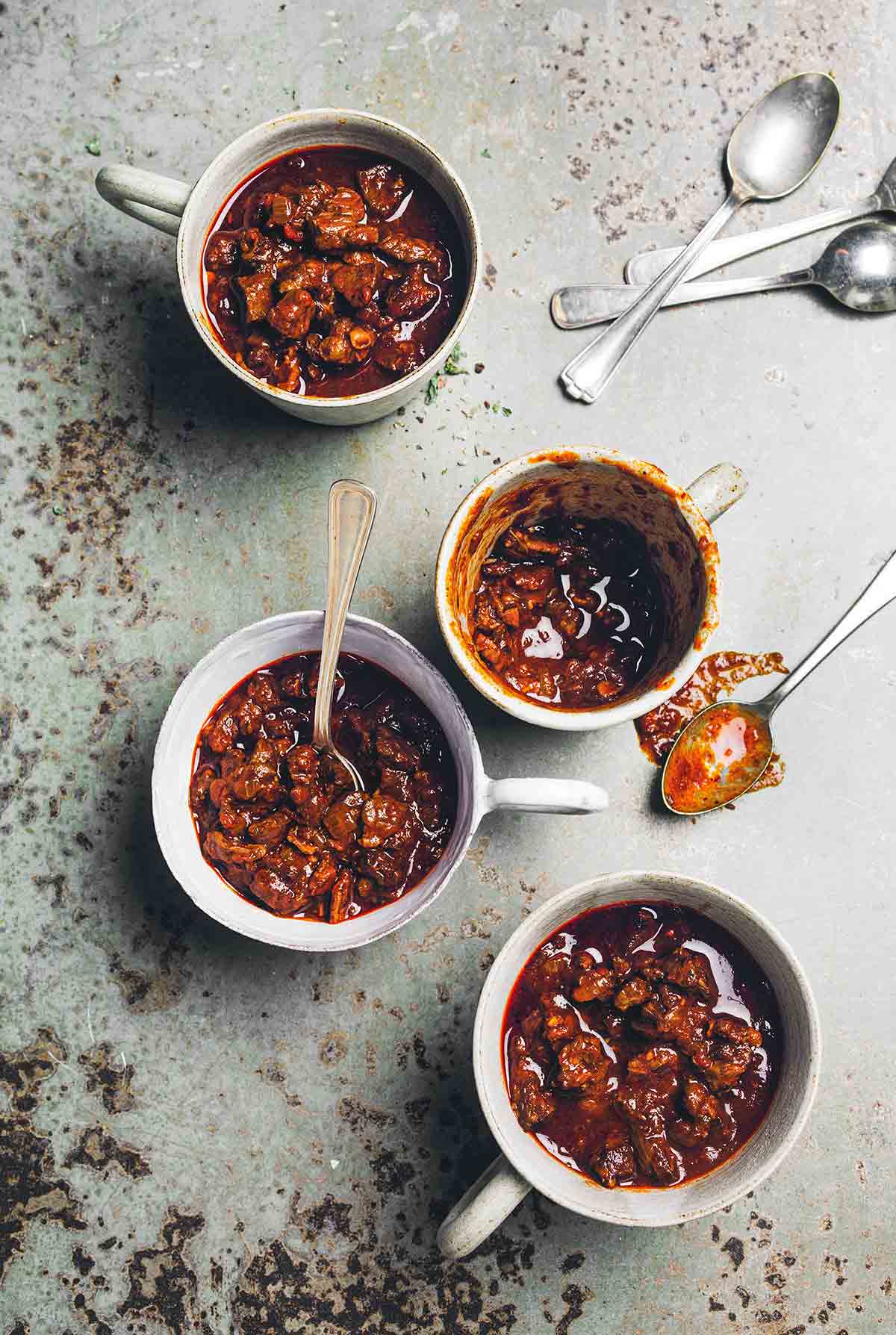 Four mugs filled with chili con carne with one spoon resting inside a mug and four others laying beside the mugs.