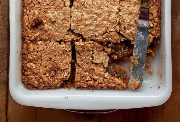 A square white baking dish filled with maple walnut squares that have been cut into 9 pieces with one missing and a knife resting in its place.