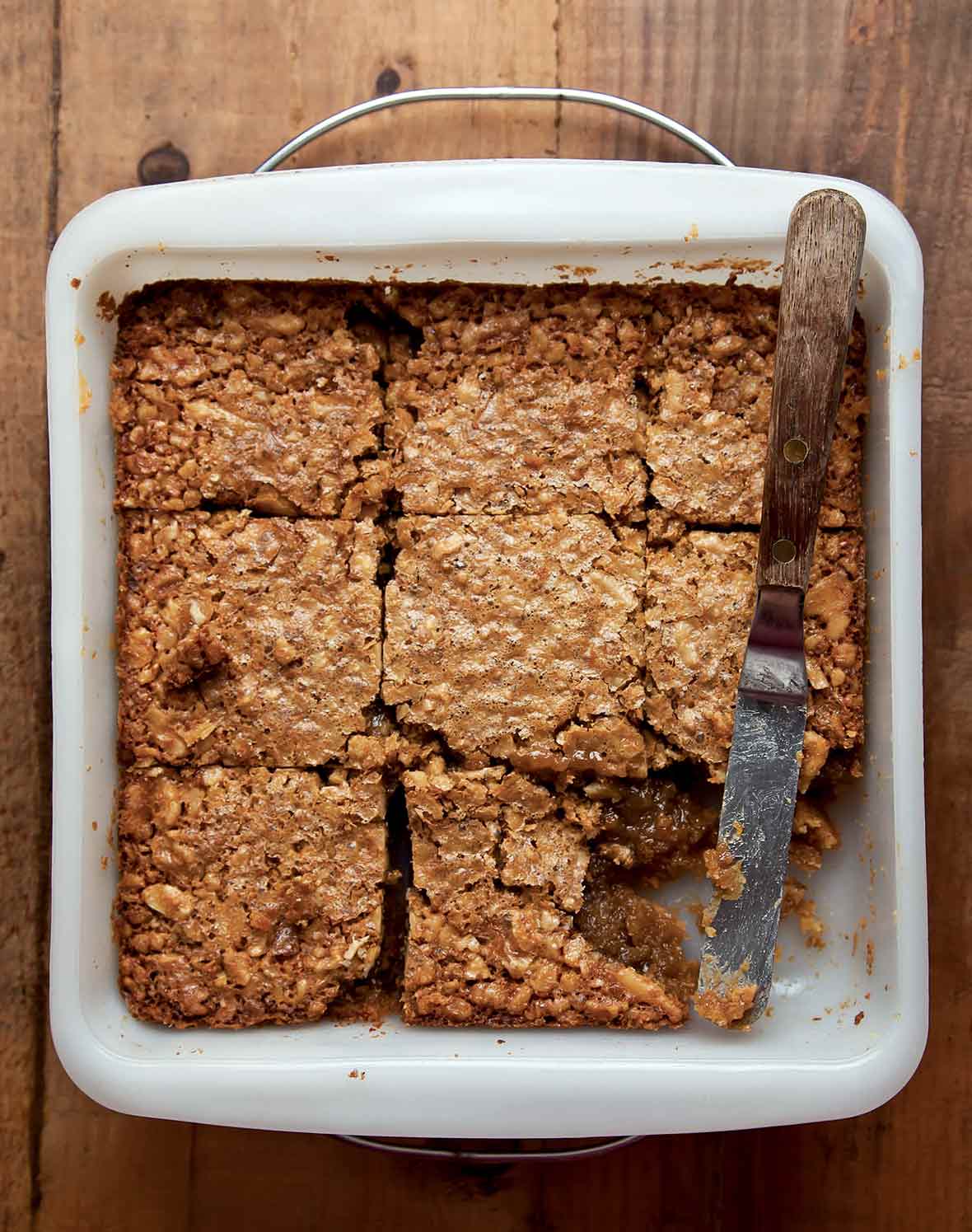 A square white baking dish filled with maple walnut squares that have been cut into 9 pieces with one missing and a knife resting in its place.