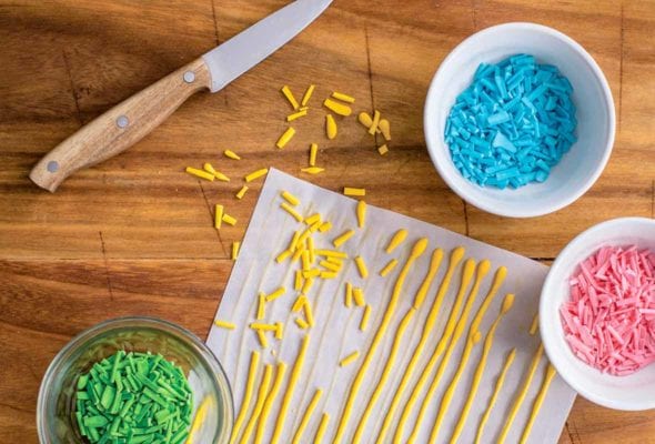 Parchment paper with long lines of yellow homemade sprinkles, plus bowls of green, blue, and pink sprinkles