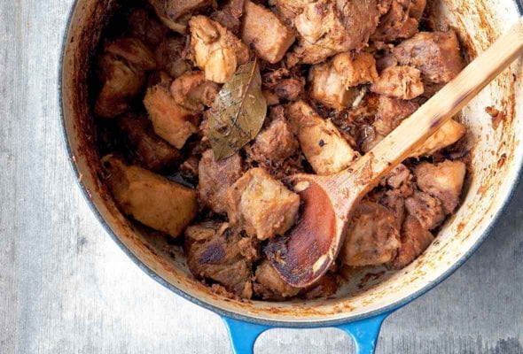 Blue pot of chunks of pork, authentic carnitas, with a bay leaf and wooden spoon on top