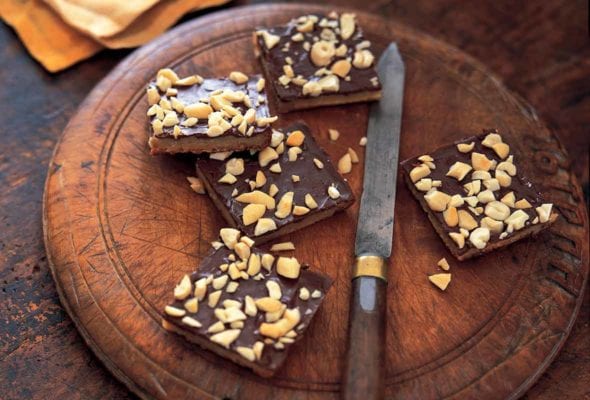 Four pieces of cashew caramel cracker bars on a round wooden board with a knife resting beside them.
