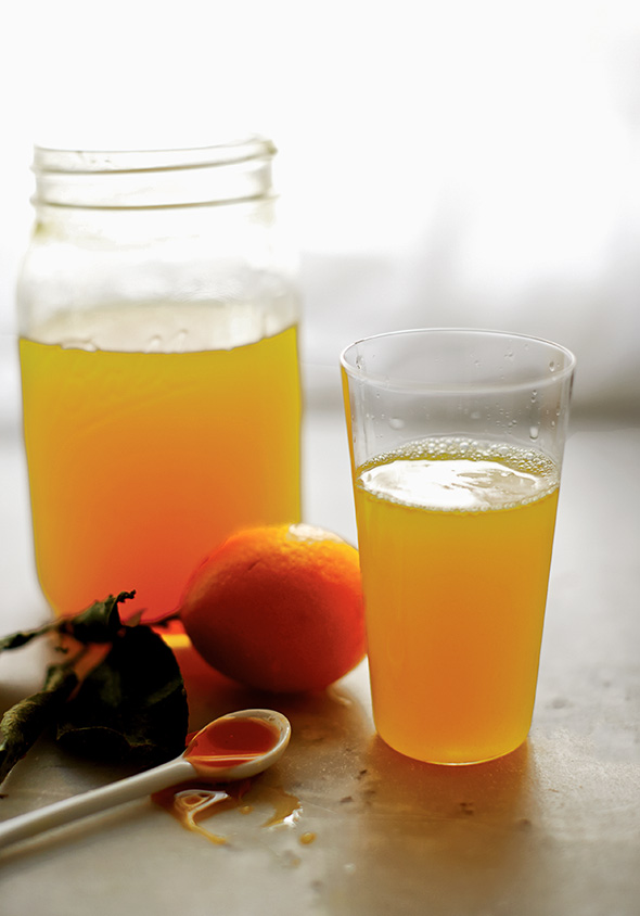 A mason jar and a tall glass filled with turmeric lemonade, and a spoon and orange lying nearby.