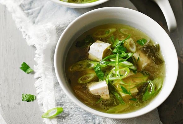 Two bowls of vegan miso soup on a grey cloth on a grey board with a spoon resting on the board.