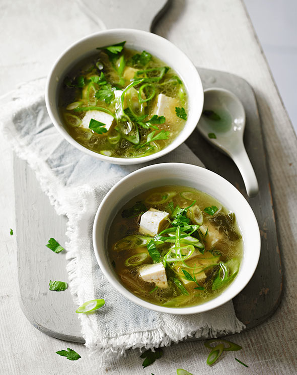 Two bowls of vegan miso soup on a grey cloth on a grey board with a spoon resting on the board.