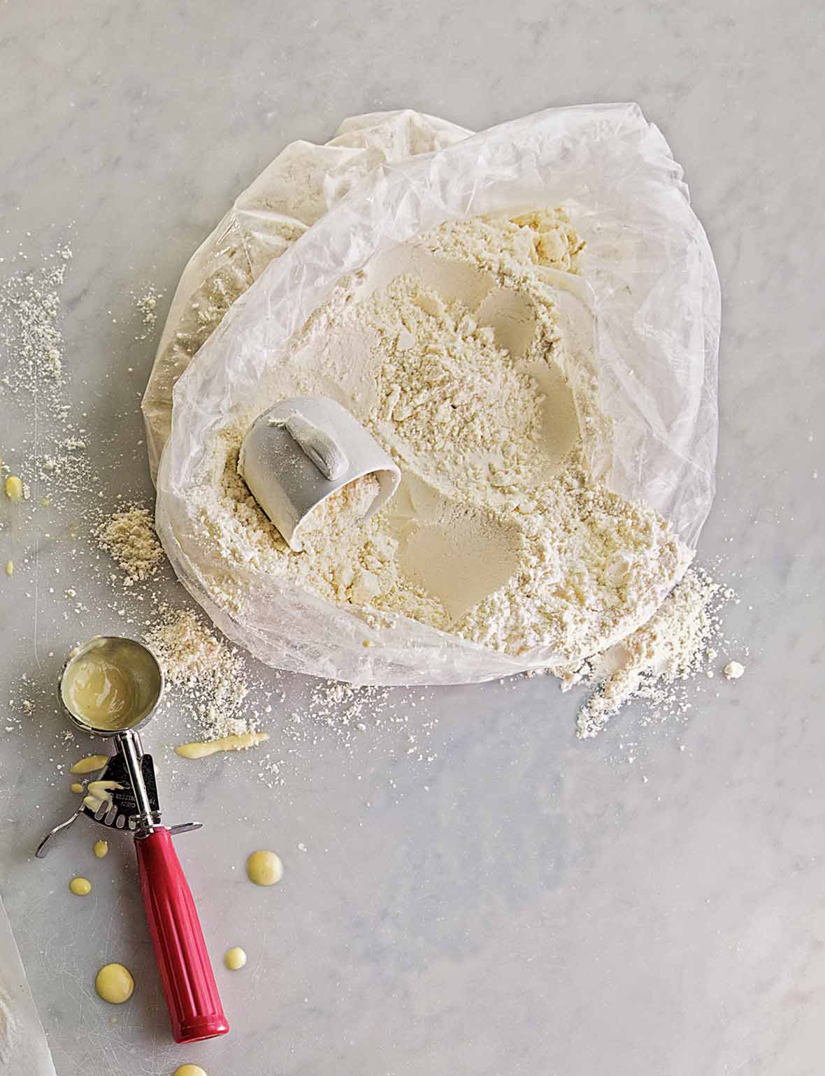 An open plastic bag of homemade pancake mix with a mug for scooping and an ice cream scoop on the side for the batter.