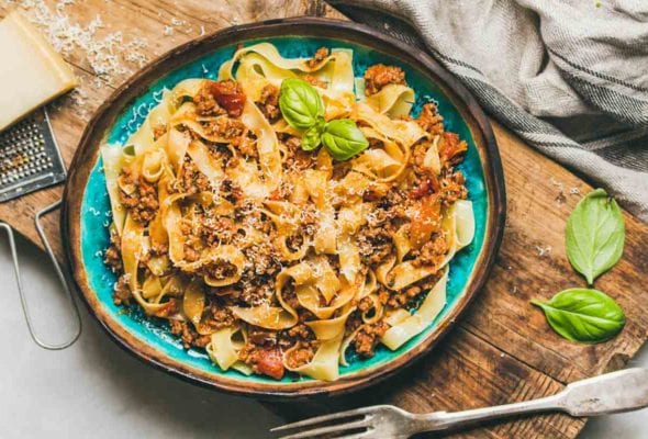 A blue bowl filled with pappardelle noodles and Marcella Hazan's bolognese sauce on a wooden board with a block of Parmesan and a grater beside the bowl.