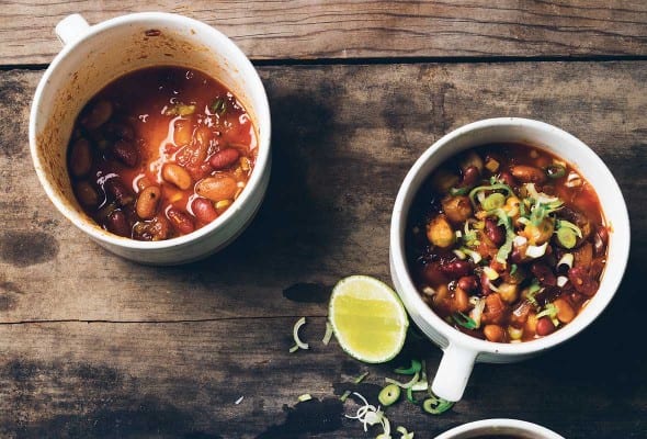Three mugs, 2 full of vegetarian chili, and one empty, with squeezed lime halves and spoons lying beside them.