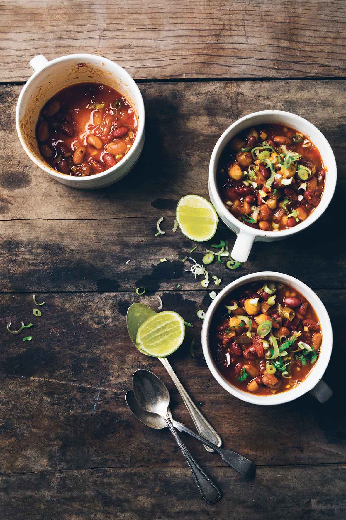 Three mugs, 2 full of vegetarian chili, and one empty, with squeezed lime halves and spoons lying beside them.