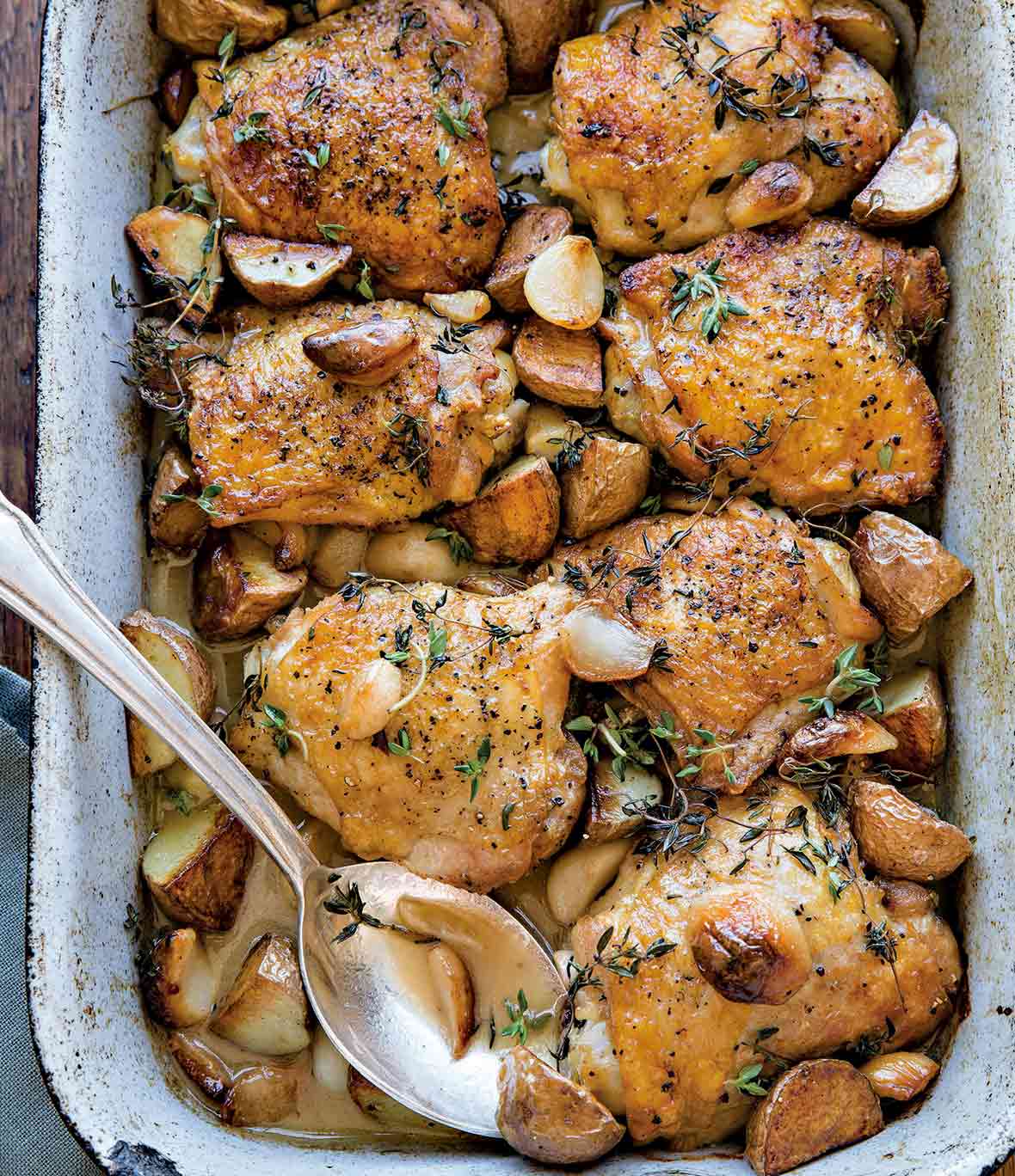 A baking dish with chicken thighs topped with garlic cloves, red potatoes, and thyme. A silver spoon is nestled in the dish.